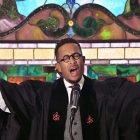 African American man delivers a sermon in front of a stained glass window.